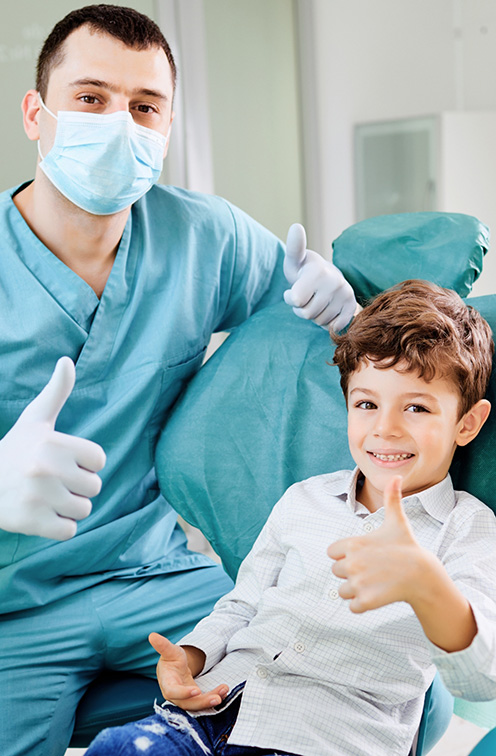 Young boy in dental chair giving thumbs up with pediatric dental team member